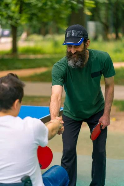 彼の手のラケットを持つ包括的な障害者男性は バックグラウンドでテニステーブルとピンポンのゲームの前に 年上の男と手を振ります — ストック写真