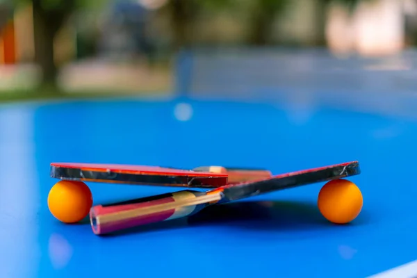 2つのテニスラケットと2つのオレンジテニスボールは 都市公園ピンポンゲームクローズアップのネットの隣にある青いテニステーブルに横たわっています — ストック写真