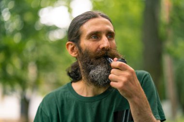 Uzun saçlı ve gri sakallı yaşlı bir adamın portresi ağaçların arkasında pipo içiyor.