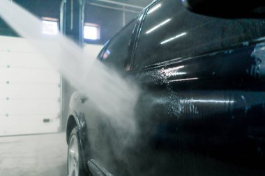 Erkek bir araba yıkama çalışanı, yüksek basınçlı çamaşır makinesiyle siyah lüks bir arabanın jantlarını yıkıyor.