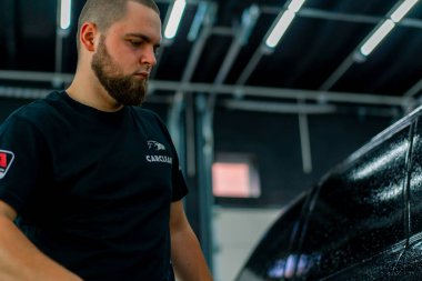 Erkek bir oto yıkama çalışanı oto yıkama kutusundaki sprey tabancayı kullanarak siyah lüks bir arabaya deterjan sürüyor.