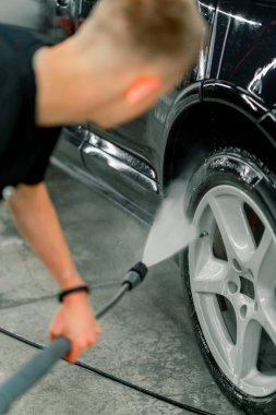 Erkek bir araba yıkama çalışanı, yüksek basınçlı çamaşır makinesiyle siyah lüks bir arabanın kenarını yıkıyor ve yoruluyor.
