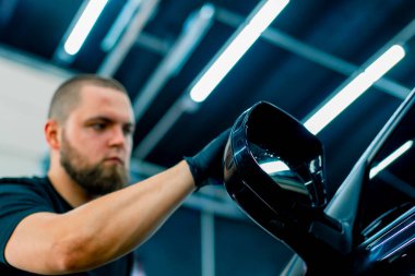 Odaklanmış erkek araba yıkama işçisi, siyah eldivenli, lüks mavi bir arabanın aynasını parlatan sünger araba bakımı konseptini kullanarak parlatıyor.