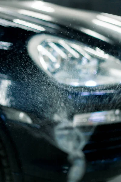 車の洗浄ボックスのスプレーボトルから洗車を適用しながら黒い車のフロントヘッドライトのクローズアップ — ストック写真