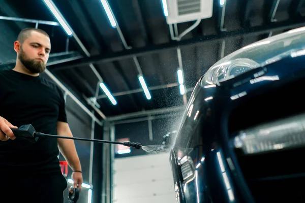 A male car wash employee applies car wash detergent to a black luxury car using spray gun in the car wash box