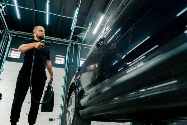 男性の洗車従業員は洗車ボックスにスプレーガンを使用して黒い高級車に洗車洗剤を適用します — ストック写真