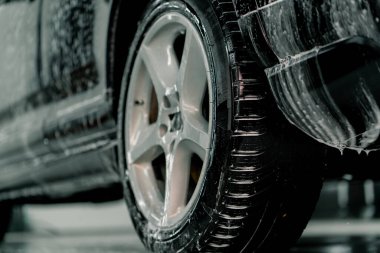 Araba bakım konseptinin araba yıkama kutusundaki siyah lüks arabanın arka lastiğine köpük uygulaması yakın plan.