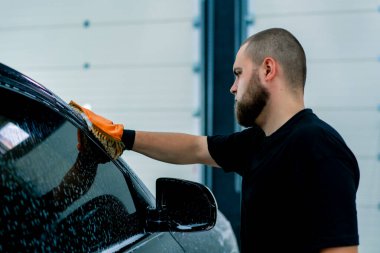 Bir oto yıkama işçisi, araba yıkama şampuanıyla siyah lüks arabanın ön farlarını yıkamak için mikrofiber bez kullanır.