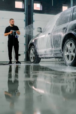 Erkek bir oto yıkama çalışanı oto yıkama kutusundaki sprey tabancayla lüks siyah bir arabaya köpük sürüyor.