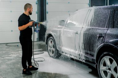 Erkek bir oto yıkama çalışanı oto yıkama kutusundaki sprey tabancayla lüks siyah bir arabaya köpük sürüyor.