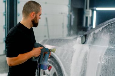 Erkek bir oto yıkama çalışanı oto yıkama kutusundaki sprey tabancayla lüks siyah bir arabaya köpük sürüyor.