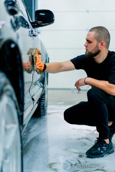 車の洗濯機の労働者は車の洗浄シャンプーが付いている黒い高級車のフロント ヘッドライトを洗浄するのにマイクロファイバーの布を使用します — ストック写真