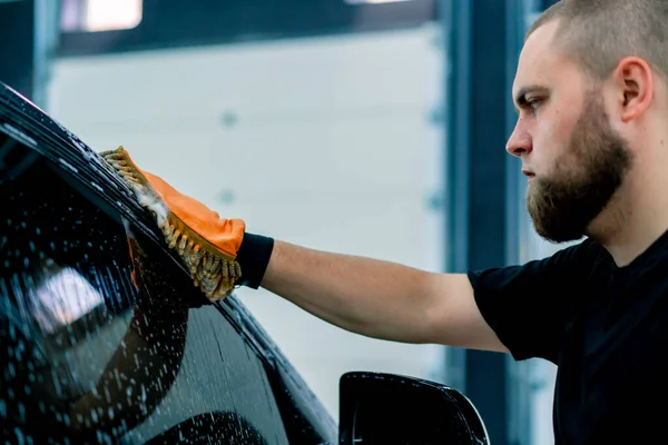 車の洗濯機の労働者は車の洗浄シャンプーが付いている黒い高級車のフロント ヘッドライトを洗浄するのにマイクロファイバーの布を使用します — ストック写真