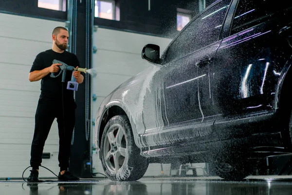 男性の洗車従業員は洗車ボックスにスプレーガンを使用して高級黒い車に洗車フォームを適用します — ストック写真
