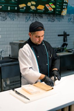 Siyah eldiven ve kep kesimi takan bir suşi şefi suşi restoranının mutfak masasında makarna, pilav ve somon balığı ile hazır.