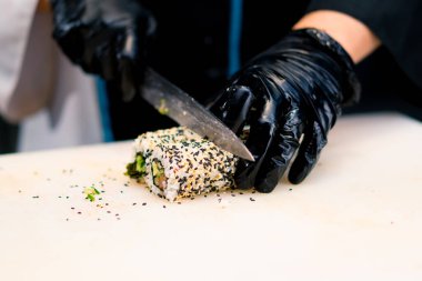 Sushi restoranının mutfağında bıçakla susamlı alabalıkla pişirdiği California dürümünü kesen bir suşi şefinin yakın çekimi.