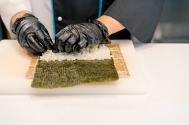 Siyah eldivenli suşi makinesinin yakın plan çekimi. Suşi hazırlamak için mutfak masasında pirinç nori yaprağı somon peynirli salatalık peyniri avokadosu var.