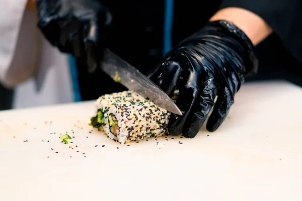 Close-up of a sushi chef cutting a california roll he cooked with trout in sesame seeds with a knife in the kitchen of sushi restaurant