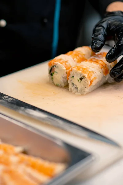 プロのキッチンで白い台所板にカリフォルニアのロールを切る黒い手袋の寿司の男のクローズアップ — ストック写真