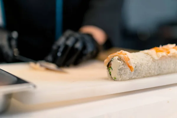 キッチンでカリフォルニアロールを準備しながらエビのフィレットをナイフで切る黒い手袋の寿司メーカーのクローズアップ — ストック写真