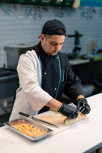 黒手袋の寿司メーカーのクローズアップ 米のシートサーモンフィレットカルドチーズキュウリのアボカドをキッチンテーブルに使用して寿司を準備 — ストック写真