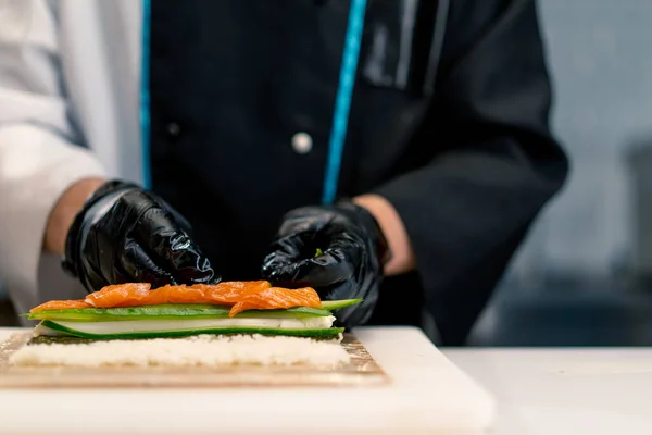 黒手袋の寿司メーカーのクローズアップ 米のシートサーモンフィレットカルドチーズキュウリのアボカドをキッチンテーブルに使用して寿司を準備 — ストック写真