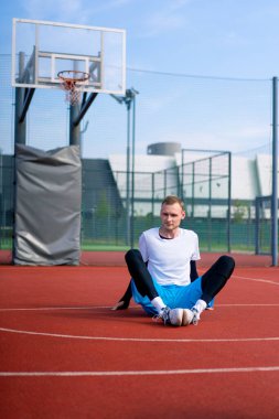 Uzun boylu basketbolcu antrenman başlamadan önce dışarıda bir basketbol sahasında oturur.