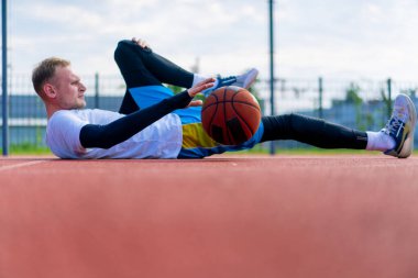 Uzun boylu basketbol oyuncusu dışarıda bir basketbol sahasında yatıyor ve antrenman başlamadan önce kaslarını esnetiyor ve topu dolduruyor.