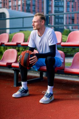 Uzun boylu basketbol oyuncusu antrenman başlamadan önce elinde topla açık hava basketbol sahasının tribünlerinde oturuyor.
