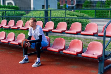Uzun boylu basketbol oyuncusu antrenman başlamadan önce elinde topla açık hava basketbol sahasının tribünlerinde oturuyor.
