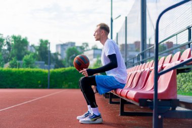 Uzun boylu basketbol oyuncusu antrenman başlamadan önce elinde topla açık hava basketbol sahasının tribünlerinde oturuyor.