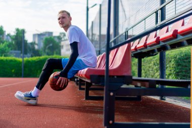 Uzun boylu bir basketbol oyuncusu açık hava basketbol sahasının tribünlerinde oturur ve antrenman başlamadan önce top sürme gösterisi yapar.