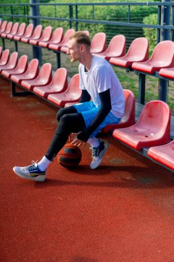 Uzun boylu bir basketbol oyuncusu açık hava basketbol sahasının tribünlerinde oturur ve antrenman başlamadan önce top sürme gösterisi yapar.