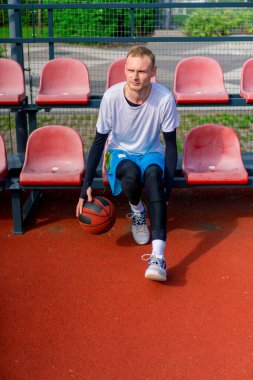 Uzun boylu bir basketbol oyuncusu açık hava basketbol sahasının tribünlerinde oturur ve antrenman başlamadan önce top sürme gösterisi yapar.