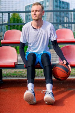 Uzun boylu bir basketbol oyuncusu açık hava basketbol sahasının tribünlerinde oturur ve antrenman başlamadan önce top sürme gösterisi yapar.