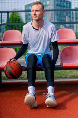 Uzun boylu bir basketbol oyuncusu açık hava basketbol sahasının tribünlerinde oturur ve antrenman başlamadan önce top sürme gösterisi yapar.