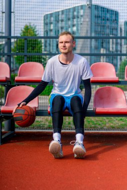 Uzun boylu bir basketbol oyuncusu açık hava basketbol sahasının tribünlerinde oturur ve antrenman başlamadan önce top sürme gösterisi yapar.
