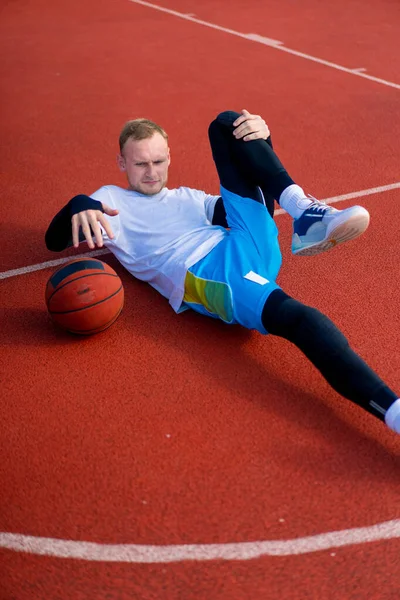 背の高い男バスケットボールプレーヤーは外のバスケットボールコートに横たわり 練習が始まる前に筋肉やボールを伸ばします — ストック写真