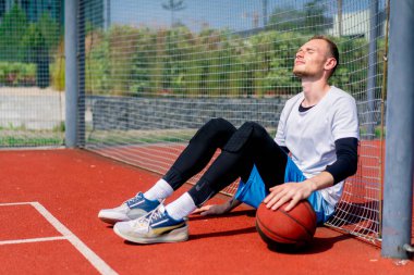 Elinde top olan uzun boylu bir basketbolcu parkta antrenman yaparken bir basketbol sahasının ağına yaslanır.