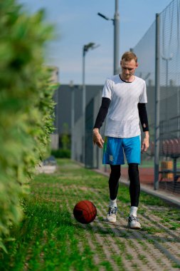 Uzun boylu basketbolcu, parktan basketbol sahasına doğru yürüyor ve topu sürüş yeteneklerini sergileyerek sürüyor.