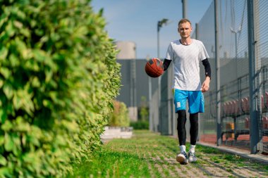 Uzun boylu basketbolcu, parktan basketbol sahasına doğru yürüyor ve topu sürüş yeteneklerini sergileyerek sürüyor.