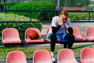 Basketbol sahasının tribünlerinde oturan uzun ve sinirli basketbol oyuncusu.