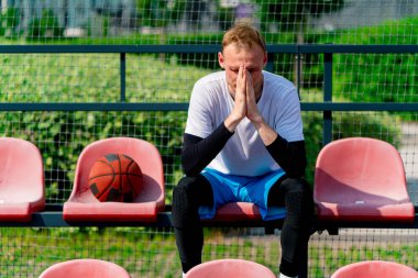 Basketbol sahasının tribünlerinde oturan uzun ve sinirli basketbol oyuncusu.