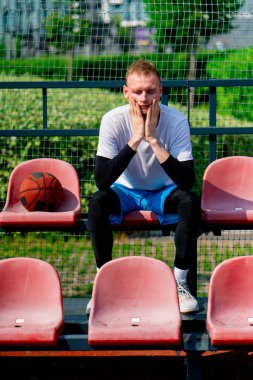 Basketbol sahasının tribünlerinde oturan uzun ve sinirli basketbol oyuncusu.