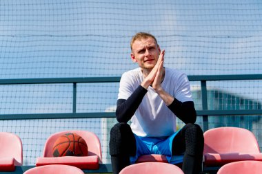 Basketbol sahasının tribünlerinde oturan uzun ve sinirli basketbol oyuncusu.