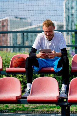 Uzun boylu bir basketbolcu, yanında topla tribünde tablete bakıyor.