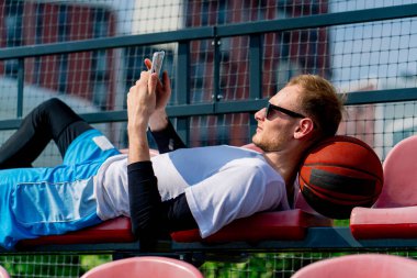 Uzun boylu basketbolcu kafasını topun üzerine koyar ve basketbol sahasının tribünlerinde bir panoya bakar.