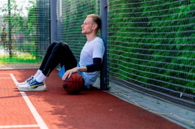 Elinde top olan uzun boylu bir basketbolcu parkta antrenman yaparken bir basketbol sahasının ağına yaslanır.