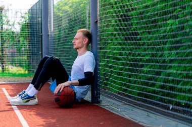 Elinde top olan uzun boylu bir basketbolcu parkta antrenman yaparken bir basketbol sahasının ağına yaslanır.