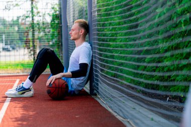 Elinde top olan uzun boylu bir basketbolcu parkta antrenman yaparken bir basketbol sahasının ağına yaslanır.